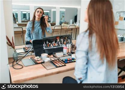 Beautiful woman with brush in hand sitting against mirror in beauty studio. Cosmetic salon