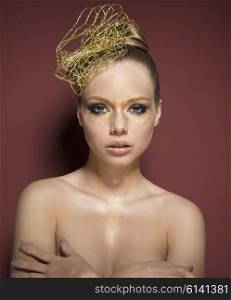 beautiful woman with creative golden glitter make-up and accessory in the hair-style. She is covering her nude breast and looking in camera