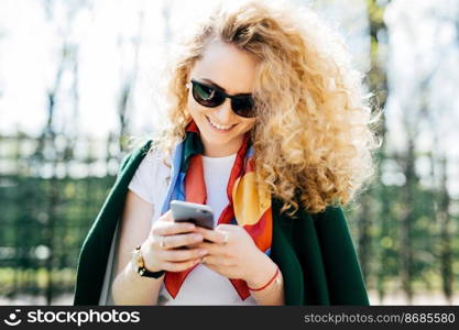 Beautiful woman with curly hair wearing sunglasses and green jacket holding smart phone in her hands browsing internet enjoying online communication while standing against green park background