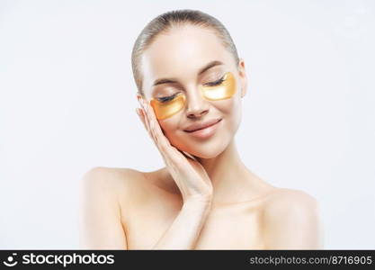 Beautiful woman with dark hair, closes eyes and touches face, applies golden collagen patches, has lifting anti wrinkle mask, stands naked shoulders, stands over white background. Spa treatment