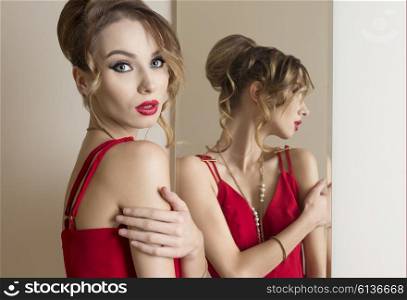 beautiful woman with elegant hairdo and make-up posing with red dress near the mirror and looking in camera