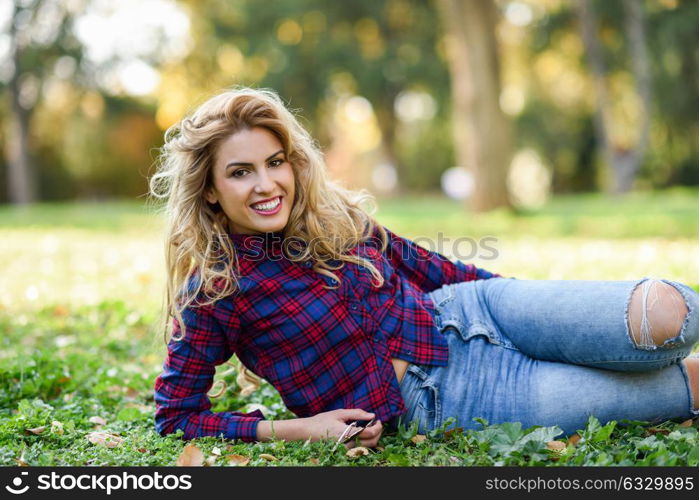 Beautiful woman with long blond curly hair smiling on the grass of a urban park. Expressive Woman in checkered shirt and blue jeans with toothy smile