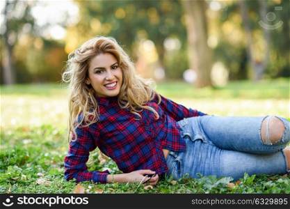 Beautiful woman with long blond curly hair smiling on the grass of a urban park. Expressive Woman in checkered shirt and blue jeans with toothy smile
