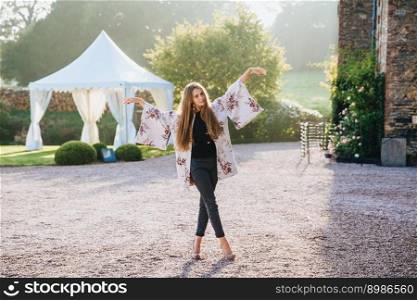 Beautiful woman with long straight hair wears white cape, jeans and high heeled shoes stands crossed legs, gestures with hands aside, poses at camera against ancient city s background, charming nature