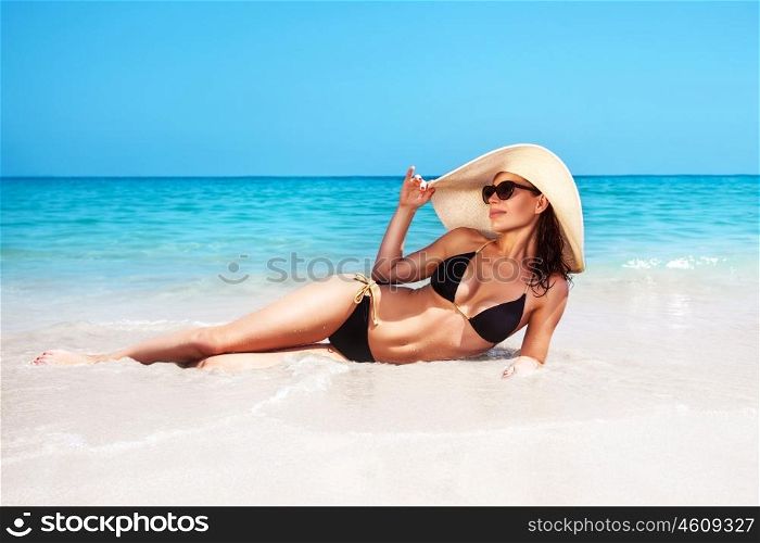 Beautiful woman with perfect body lying down on the beach, wearing stylish hat and tanning, spending holidays on a tropical beach resort, happy summer vacation