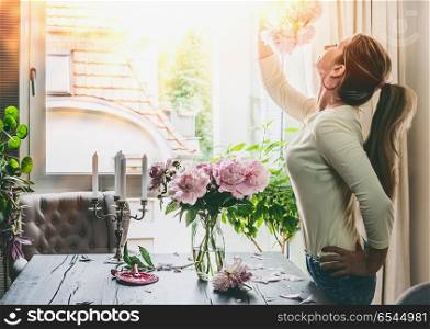 Beautiful woman with pink flowers peonies on the table in the living room composes a bouquet in a glass vase at window. Summer still life. Cozy home scene