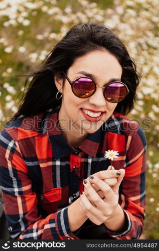 Beautiful woman with sunglasses sitting in the middle of a flowery meadow smelling a daisy