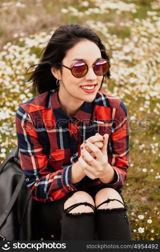 Beautiful woman with sunglasses sitting in the middle of a flowery meadow smelling a daisy