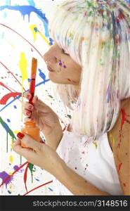 Beautiful woman with white hair and white dress covered in paint. Holding brush and bottle, thinking.