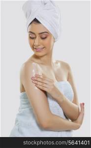 Beautiful woman wrapped in towel applying moisturizer on arm over gray background