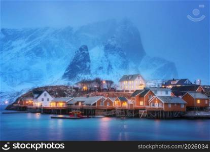 Beautiful yellow rorbuer and houses in Sarkisoy village, Lofoten islands, Norway. Winter landscape with traditional norwegian rorbuer, sea, snowy mountains in fog at night. Old fishermen&rsquo;s houses