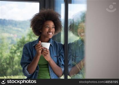 Beautiful young african american woman drinking coffee and looking through a window in her luxury home
