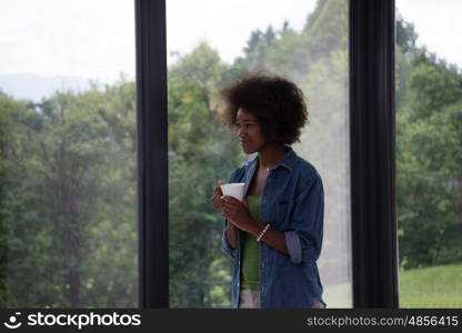 Beautiful young african american woman drinking coffee and looking through a window in her luxury home