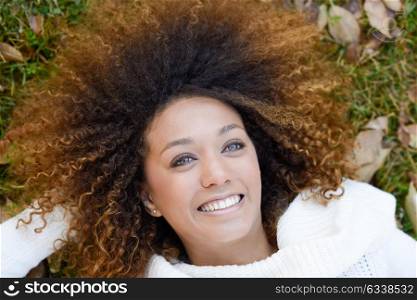 Beautiful young African American woman smiling with afro hairstyle and green eyes wearing white winter dress laying on the grass of an urban park