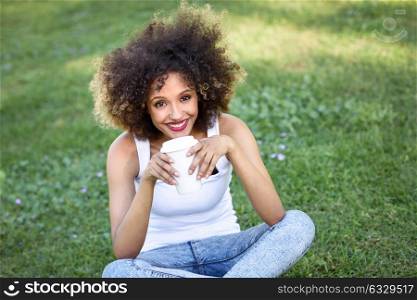 Beautiful young African American woman with afro hairstyle. Girl drinking coffee in park sitting on grass wearing casual clothes smiling.