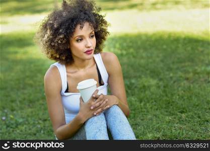 Beautiful young African American woman with afro hairstyle. Girl drinking coffee with a take away glass in park sitting on grass wearing casual clothes smiling.
