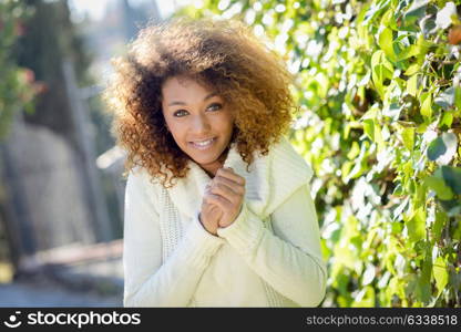 Beautiful young African American woman with afro hairstyle and green eyes wearing white winter dress with autumn leaves in the background.