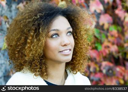 Beautiful young African American woman with afro hairstyle and green eyes wearing white winter dress with autumn leaves in the background.
