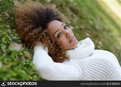 Beautiful young African American woman with afro hairstyle and green eyes wearing white winter dress laying on the grass of an urban park