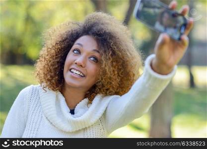 Beautiful young African American woman with afro hairstyle and green eyes wearing white winter dress. Beautiful Girl selfie in the park with a smartphone.