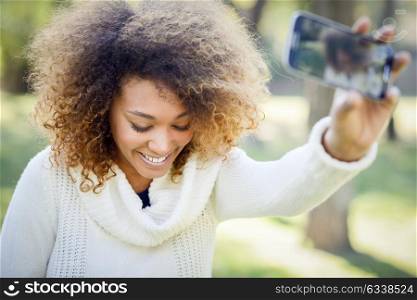 Beautiful young African American woman with afro hairstyle and green eyes wearing white winter dress. Beautiful Girl selfie in the park with a smartphone.
