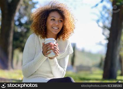 Beautiful young African American woman with afro hairstyle and green eyes wearing white winter dress. Girl drinking coffee in park sitting on grass wearing casual clothes smiling.