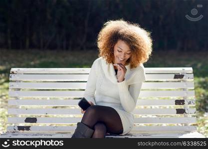 Beautiful young African American woman with afro hairstyle wearing white winter dress. Beautiful Girl sitting in the park with a smartphone.
