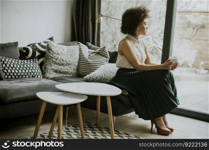 Beautiful young afro american young woman with curly hair sitting by the window and holding a cup of coffee