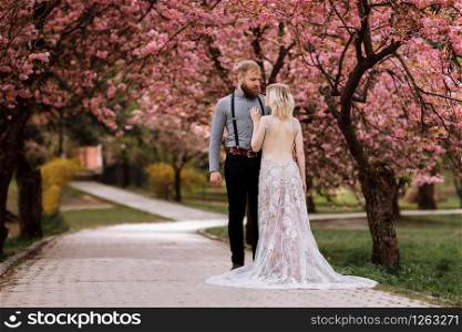 Beautiful, young and cute woman in luxury dress and bearded hipster man in blossoming pink cherry or sakura garden, hugging and looking to each other on a sunny day. Spring wedding portrait.. Beautiful, young and cute woman in luxury dress and bearded hipster man in blossoming pink cherry or sakura garden, hugging and looking to each other on a sunny day. Spring wedding portrait