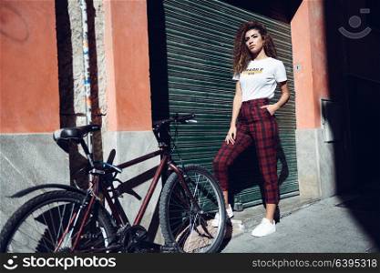 Beautiful young arabic woman with black curly hairstyle. Arab girl in casual clothes in the street near a bicycle. Girl power concept.