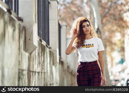 Beautiful young arabic woman with black curly hairstyle. Arab girl in casual clothes in the street. Happy female wearing white t-shirt and checked pants.