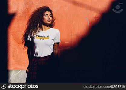 Beautiful young arabic woman with black curly hairstyle to the sun with her eyes closed. Arab girl in casual clothes in the street. Girl power concept.