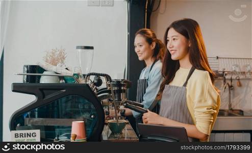 Beautiful young Asia lady barista working with coffee machine in coffee shop. Two small business owner Korean girl in apron making coffee by coffee machine with friend at counter in urban cafe.