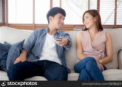 Beautiful young asian couple holding remote and watching tv or video streaming on sofa with relax and happy in living room at home, lifestyle family leisure with entertainment enjoy together on couch.