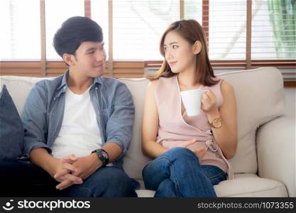 Beautiful young asian couple smiling and talking story married sitting on sofa at home together with woman holding a cup of coffee, man and woman relax and enjoy, lifestyle concept.