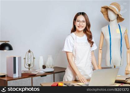 Beautiful young Asian fashion designer or costume smile and use laptop computer while design a beautiful sombrero and taking on mannequin