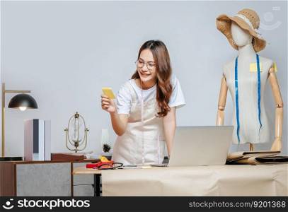 Beautiful young Asian fashion designer or tailor with working with laptop computer and read on paper note, full of tailoring tools with mannequin and equipment on the desk in the studio
