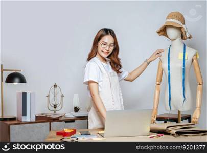 Beautiful young Asian fashion designer or tailor with working with laptop computer to search idea or online orders, full of tailoring tools  with mannequin and equipment on the desk in the studio