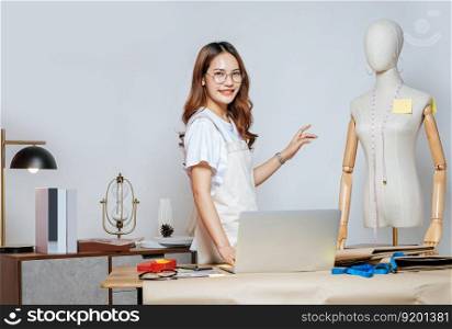Beautiful young Asian fashion designer or tailor with working with laptop computer to search idea or online orders, full of tailoring tools with mannequin and equipment on the desk in the studio