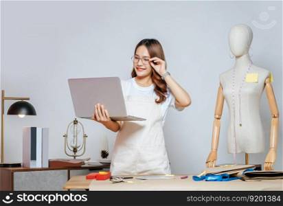 Beautiful young Asian fashion designer or tailor with working with laptop computer to search idea or online orders,  full of tailoring tools and equipment on the desk in the studio