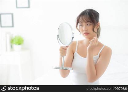 Beautiful young asian woman looking the mirror with acne problem at bedroom, trouble of beauty on face, zit treatment, asia girl is pimple having worry and displeased, skincare and healthy concept.