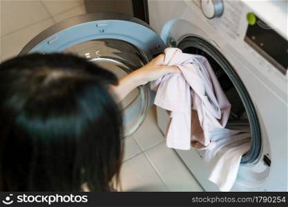 Beautiful young Asian woman putting clothes into washing machine in kitchen at home. Laundry concept. Closeup.