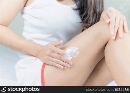 Beautiful young asian woman sitting on a bed stroking legs with soft smooth skin in the bedroom, girl applying touch body cream and lotion with treatment care concept.
