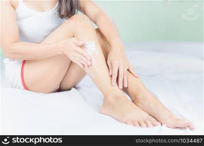 Beautiful young asian woman sitting on a bed stroking legs with soft smooth skin in the bedroom, girl applying touch body cream and lotion with treatment care concept.