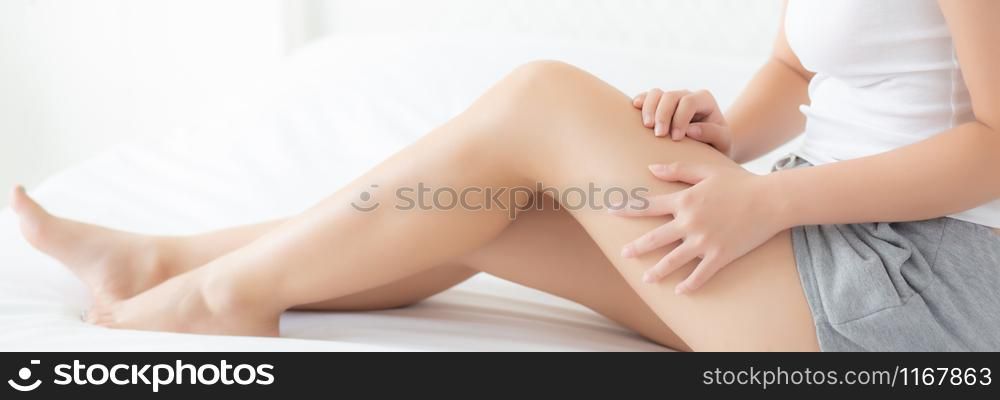 Beautiful young asian woman sitting on a bed stroking legs with soft smooth skin in the bedroom, girl applying touch body cream and lotion with treatment care, healthy and wellness concept.