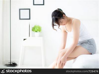 Beautiful young asian woman sitting on a bed stroking legs with soft smooth skin in the bedroom, girl applying body cream and lotion with treatment care, healthy and wellness concept.