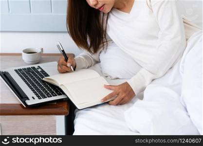 Beautiful young Asian woman sitting on bed and writing on notebook while using tablet computer, working on bed , work at home, busy lifestyle concept.