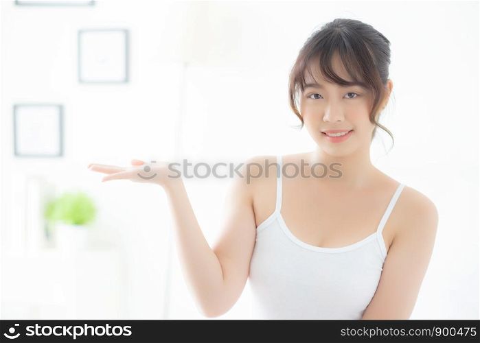 Beautiful young asian woman smiling and showing skin care product for advertisement at bedroom, beauty asia girl presenting makeup cosmetic for advertise with surprise on palms at room.