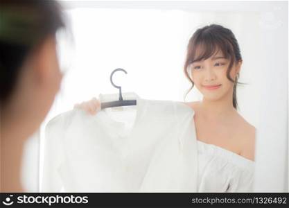 Beautiful young asian woman smiling with clothes trying on dress up fitting with modern and looking reflection mirror in the room, girl choose clothes fashion with happy and cheerful, lifestyle concept.