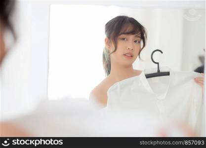 Beautiful young asian woman smiling with clothes trying on dress up fitting with modern and looking reflection mirror in the room, girl choose clothes fashion with happy and cheerful, lifestyle concept.
